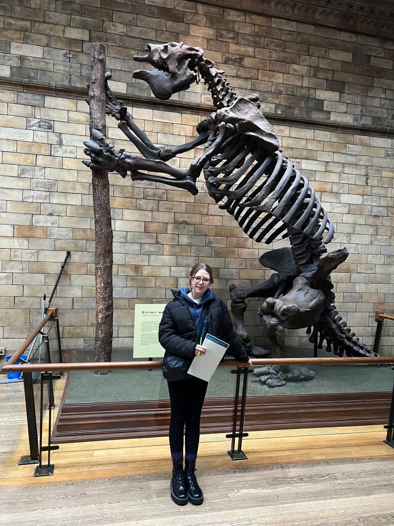 An 11 year old girl wearing black leggings, a black jacket and glasses stands in front of the skeleton f a giant sloth.