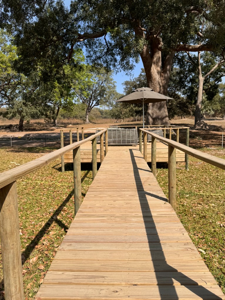 A wooden walkway with wooden railings on each side. At the end there is a small seating area with a thatched umbrella. 