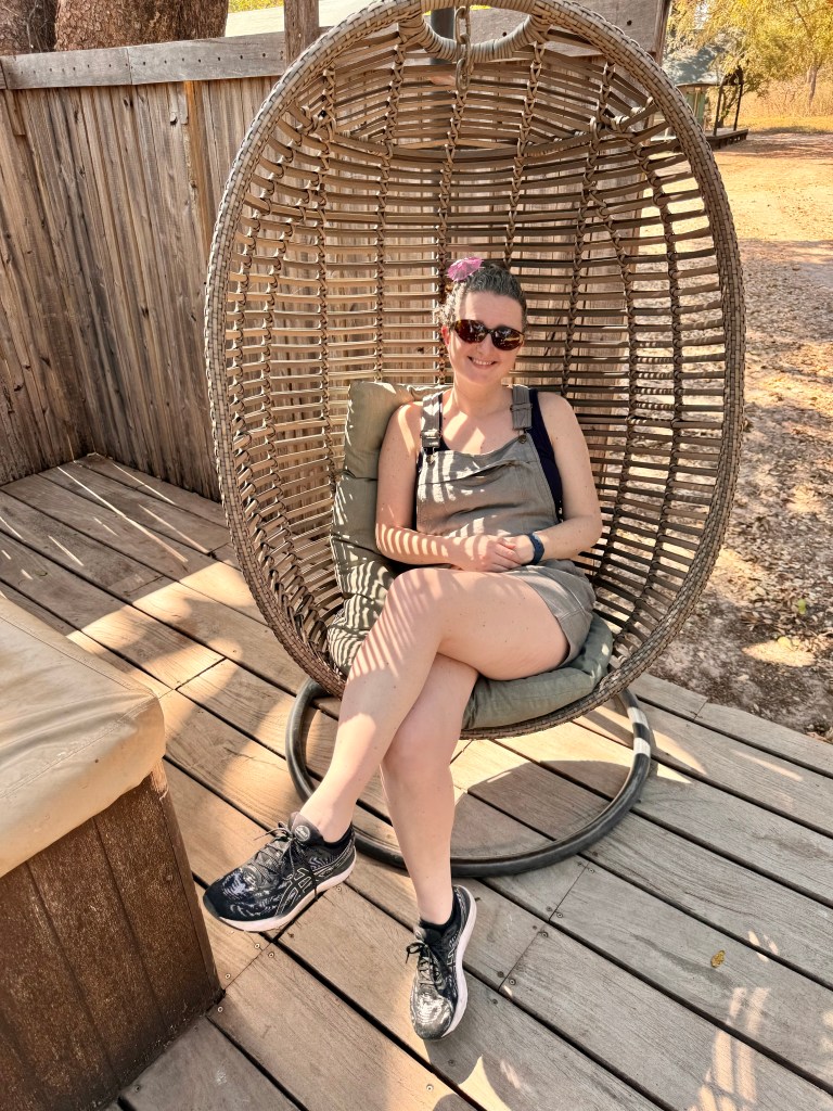 A white woman in her forties wearing khaki dungaree shorts, a black vest top and black trainers. She is sitting in a wicker egg chair on the wooden deck of the tent. 