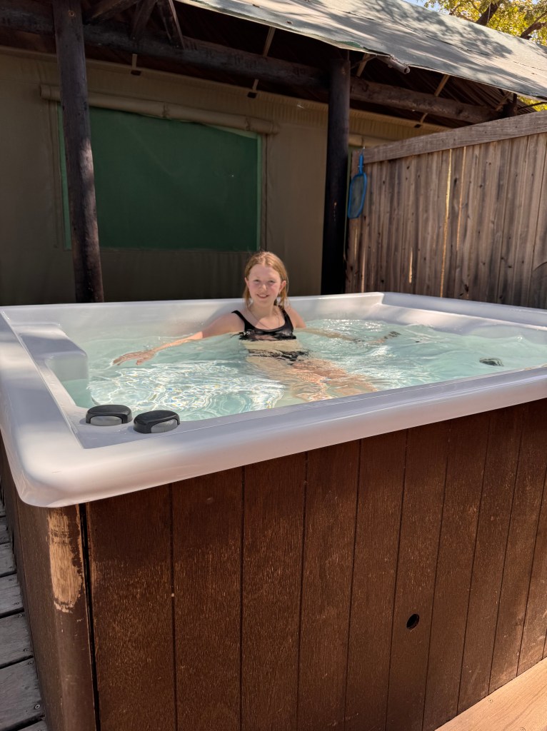 An 11 year old girl sits in a hot tub on the deck of the tent. Behind her is the tent and some wooden screening. 