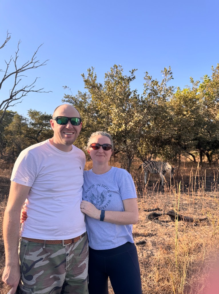 A man and a woman in their forties stood in a clearing with trees and a zebra behind them. 