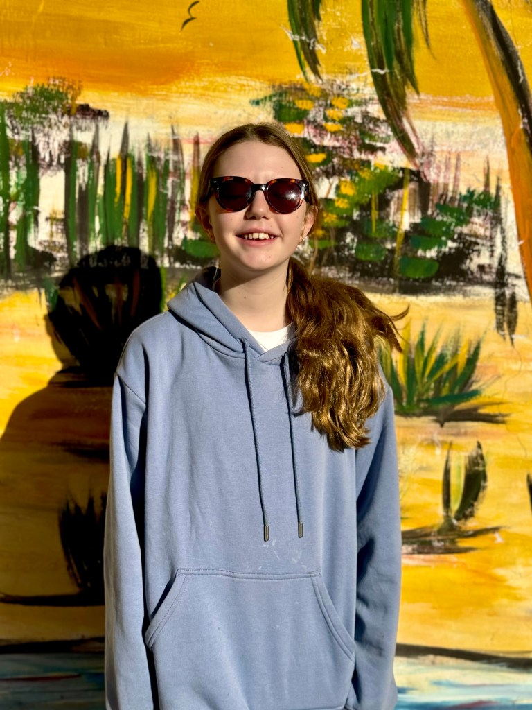 An 11 year old girl with her hair tied back in to a pony tail hanging over her shoulder and sunglasses. She is wearing a light blue hoodie and smiling at the camera. The wall behind her is painted in bright yellow with family trees and bushes. 