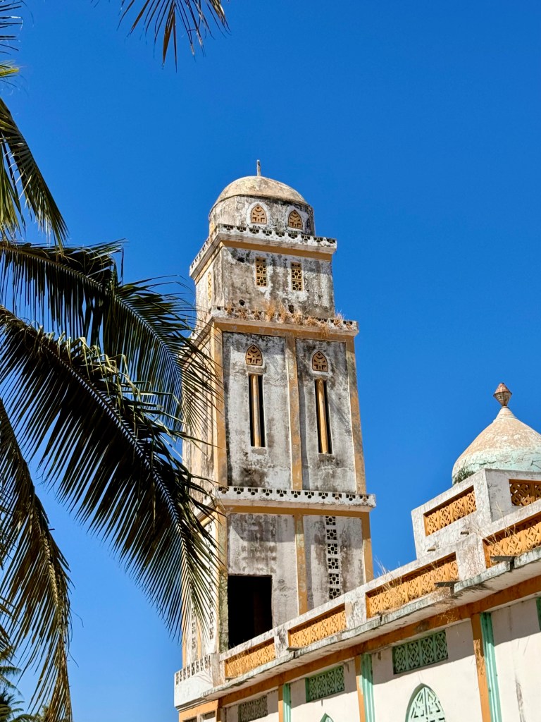 The minaret of a mosque with blue sky behind and a palm tree