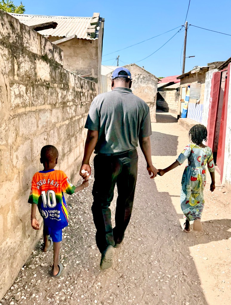 A man wearing a grey polo shirt and black trousers walks away from the camera through the streets on a Senegalese fishing village. 
He holds hands with two small children, on a left a boy wearing a brightly coloured football shirt and blue shorts and on the left a girl wearing a long floral dress. 