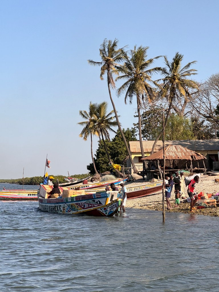 A Senegalese fishing boat moored next to a beach with fishermen unloading the contents. In the background are several palm trees. 