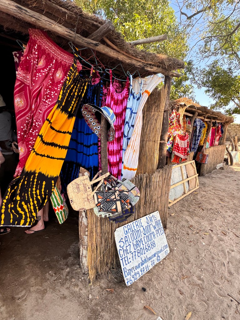 A shop in a wooden hot with various brightly coloured items of clothing hanging outside and a hand painted board with the shop details leaning against the front wall. 