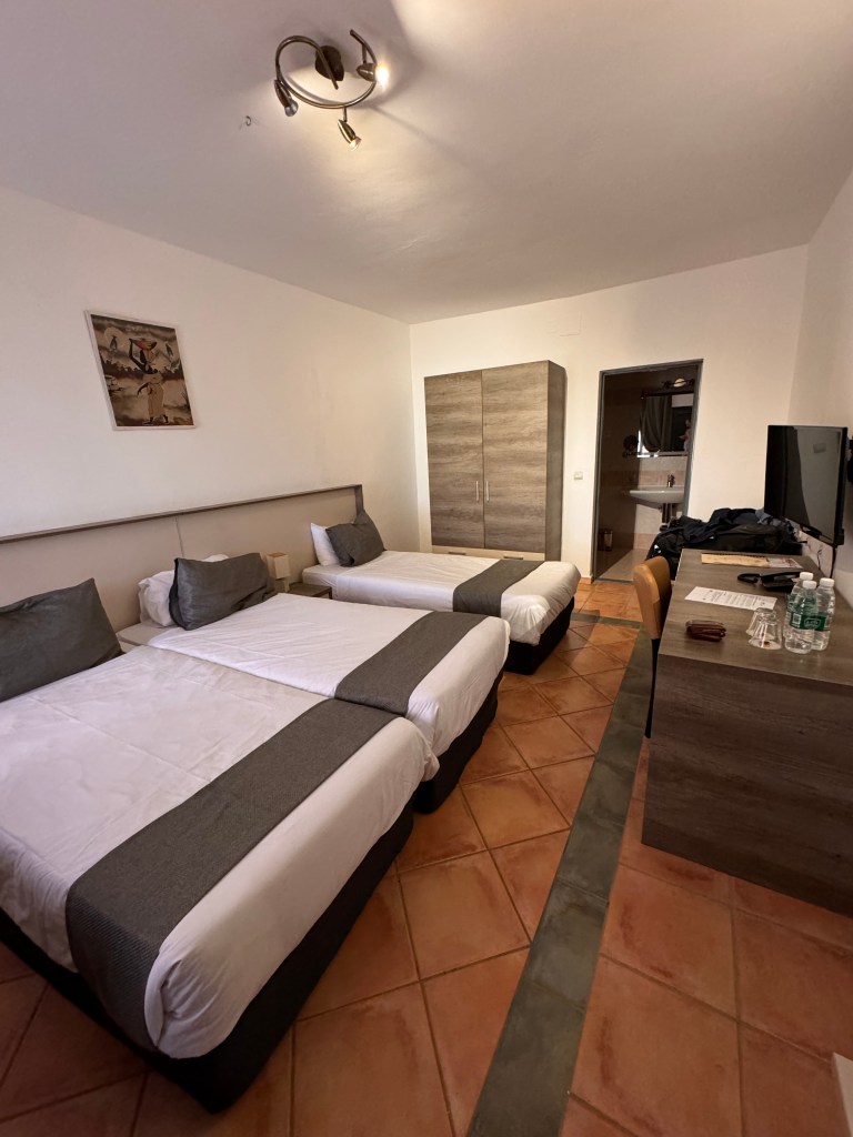 A view of the bedroom from the door showing two beds pushed together to make a double and a single. The beds are made with white sheets and a brown throw and cushion. the floor is terracotta tiles, the walls white and there is a wardrobe on the back wall and a desk on the right. 