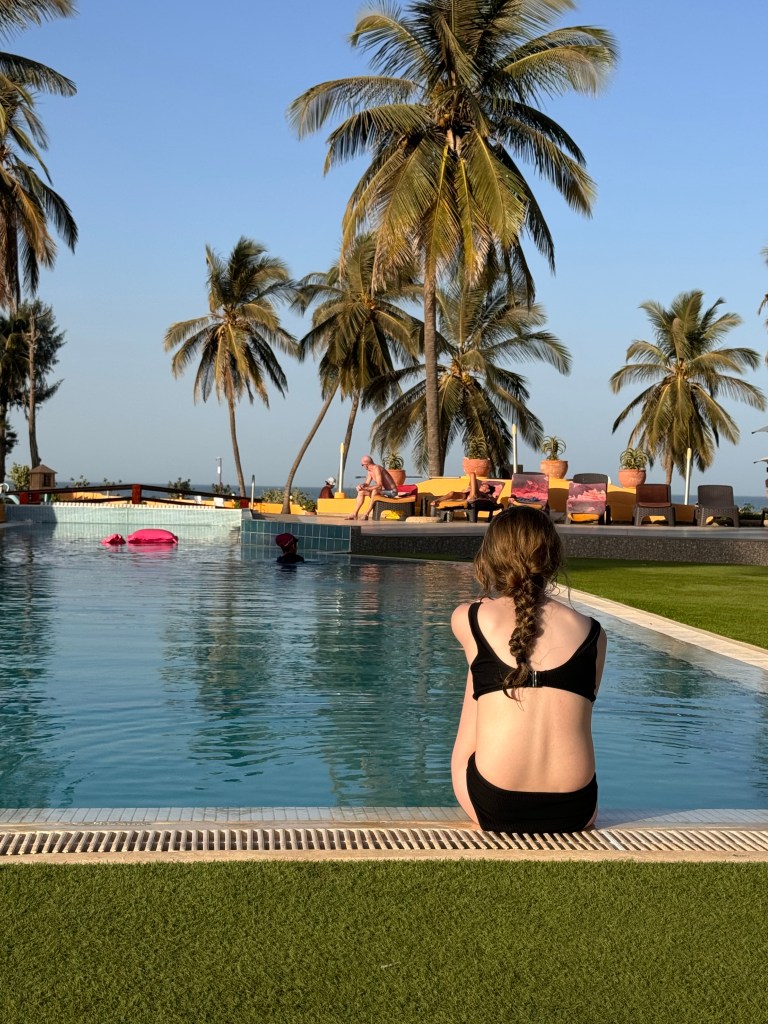 An 11 year old girl wearing a black bikini sits on the side of the pool facing away from the camera. At the other side of the pool are some sunbeams and palm trees. 