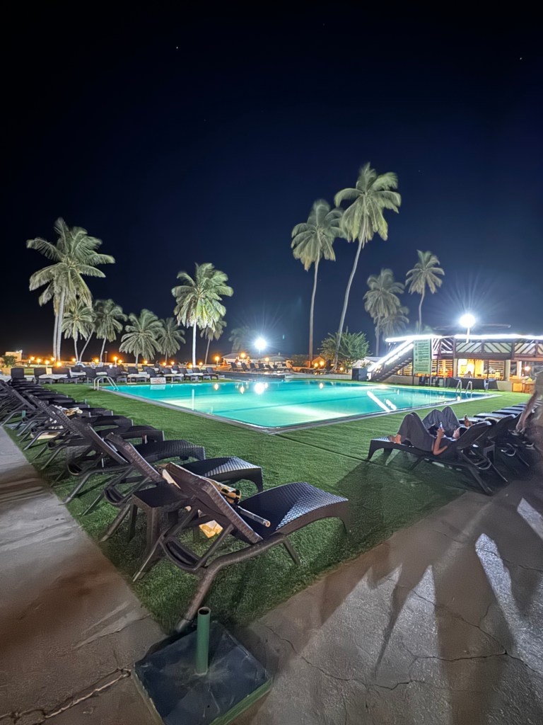 The pool area at night with the sun beds lined up around the pool and palm trees in the back ground. The pool is lit from below the water. 