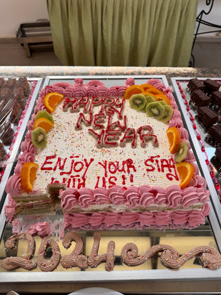 A large square cake iced with the wording 'Happy New Year, Enjoy your stay with us'. The cake is decorated with pink and white icing and kiwi and orange slices. There is a small piece of cake missing from the bottom left corner. 