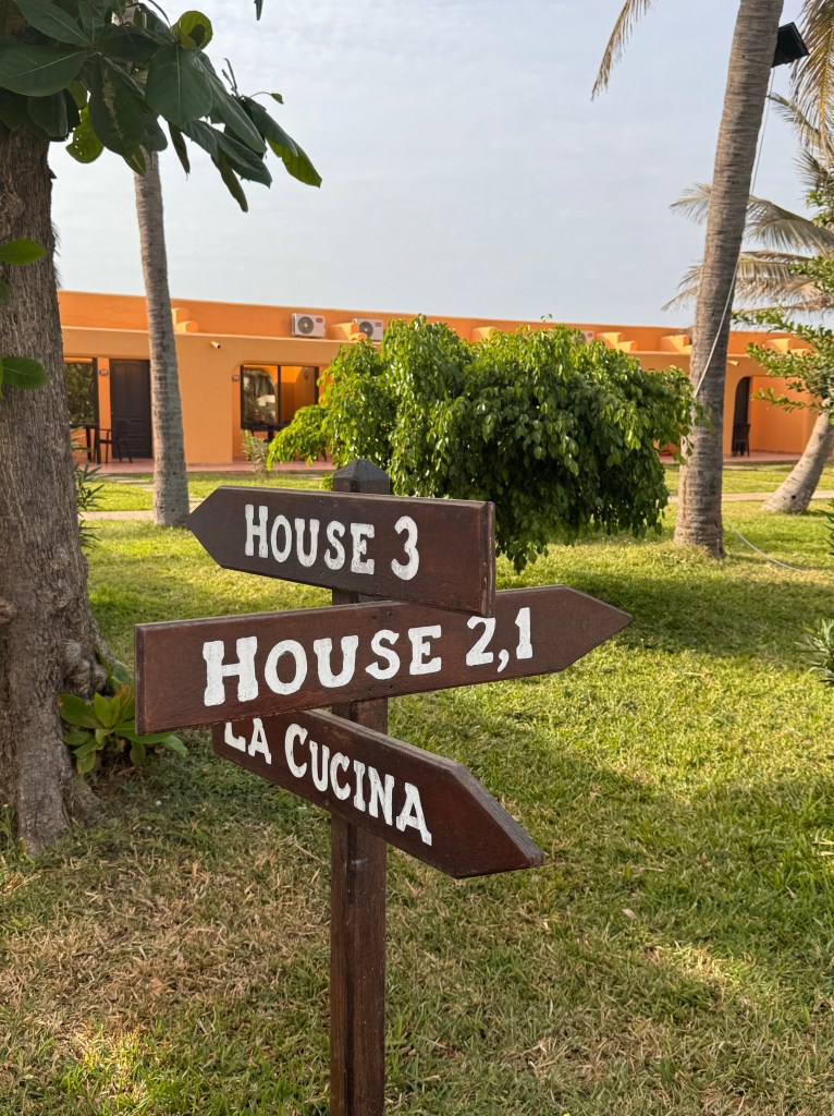 A sign post with signs for 'House 3', 'House 2, 1' and 'La Cucina'. Behind is a low yellow building and gardens and trees. 