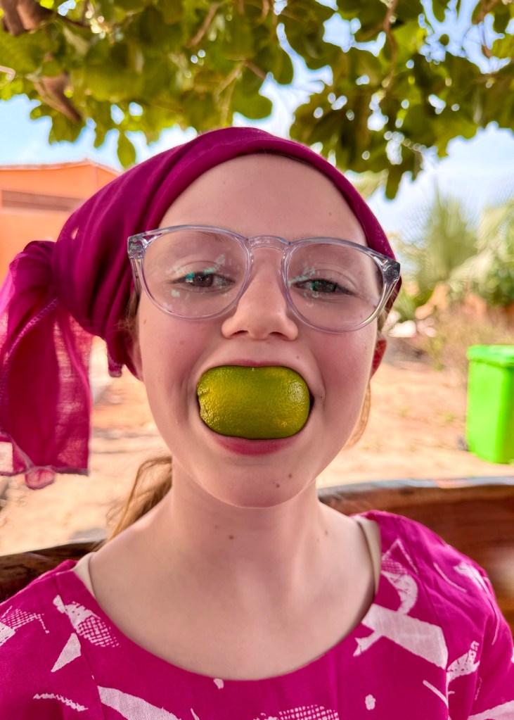 An 11 year old white girl wearing glasses and traditional Gambian dress in bright pink with a pink head wrap. She is smiling and has quarter of a green orange in her mouth. 