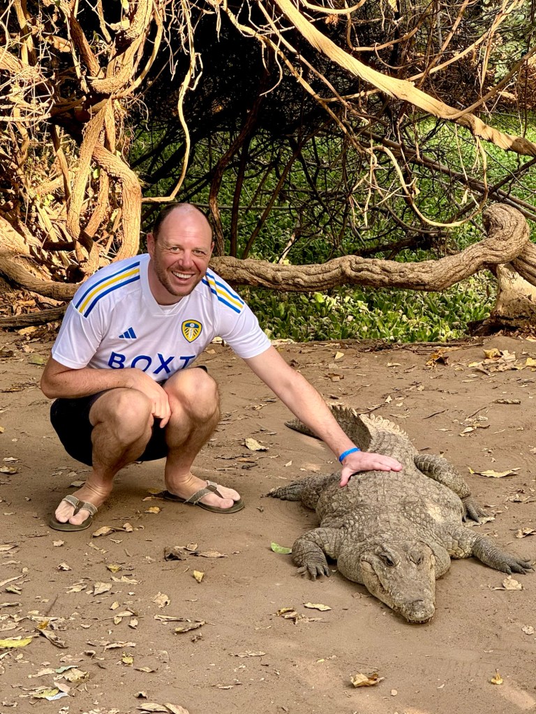 A white main his forties wearing a white Leeds United top and blue shorts is crouched down next to a crocodile with his hand on the crocodile's back. 