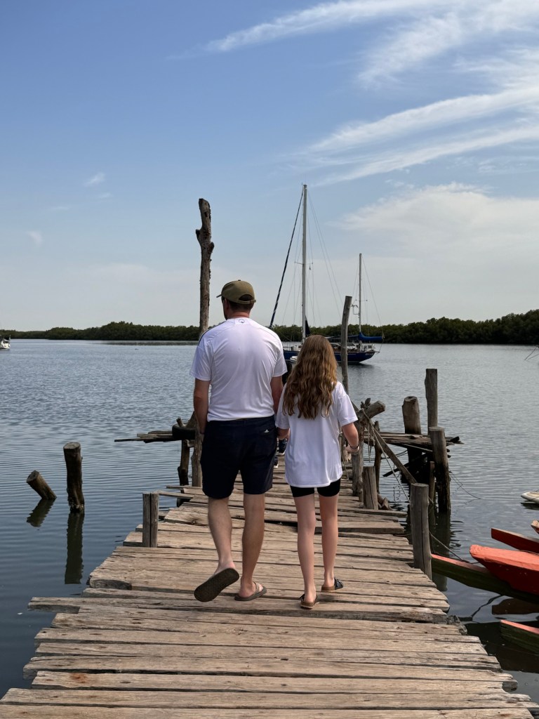 A white man in his forties wearing a white t-shirt and blue shorts and an 11 year old white girl wearing a white t-shirt and black shorts walk away from the camera down a wooden pier. 