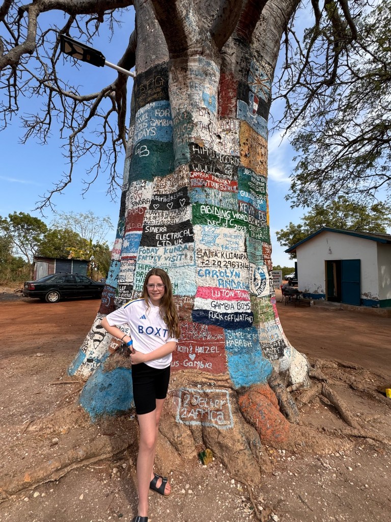 A white 11 year old girl wearing a white Leeds United shirt and black shorts is stood smiling next to a large tree trunk painted with a variety of colourful adverts. 