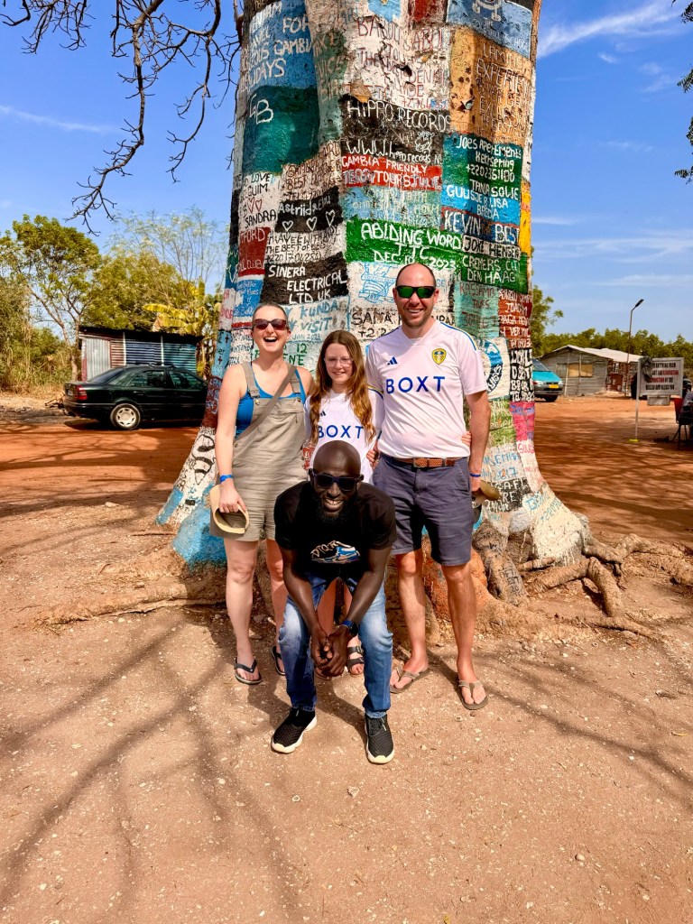 A white woman wearing sunglasses and khaki dungaree shorts, an 11 year old girl wearing a white Leeds United shirt and a white man in his forties wearing a white Leeds United shirt and blue shorts. In front of them a black man wearing jeans and a black t-shirt is crouched down. They are all in front of a large tree trunk which is painted with brightly coloured adverts. 