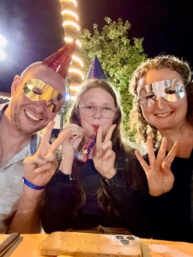 A white man in his forties wearing a light coloured shirt, a white 11 year old girl with long light brown hair and a white woman wearing a black top and with brown curly hair. They are wearing an assortment of party hats and eye masks and holding their fingers up to show the numbers 2024. 