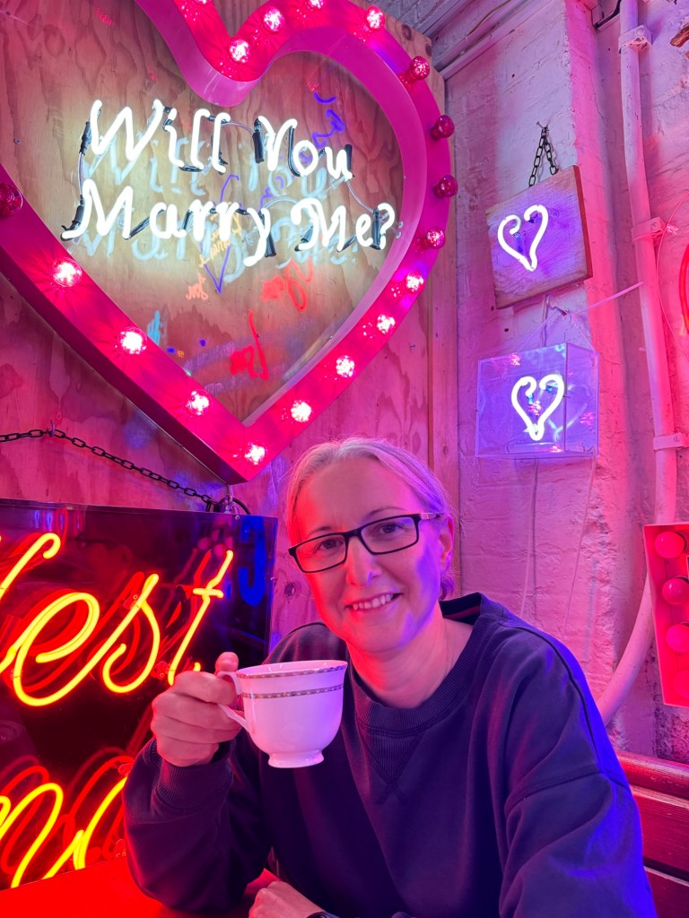 A white woman with grey hair tied back from her face and glasses sits with a cup of tea in front of a neon sign that says 'Will you marry me?'