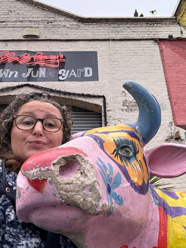 A woman in her forties with dark curly hair and glasses poses with a life-sized statue of a cow painted pink stands in front of the entrance to God's Own Junkyard. 
