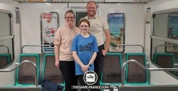 A woman wearing a pink sweatshirt, an 11 year old girl wearing a blue t-shirt and a man wearing a white t-shirt stand in a metro carriage smiling at the camera.