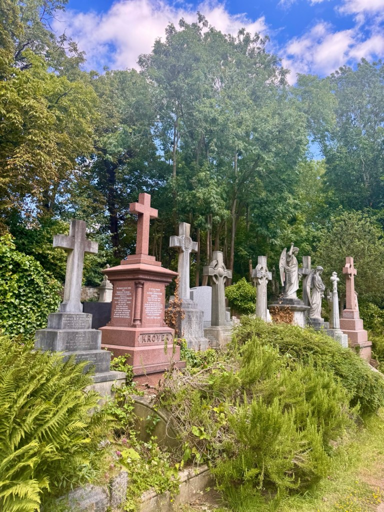 A row of gravestones in the shape of crosses amongst tress and bushes. 