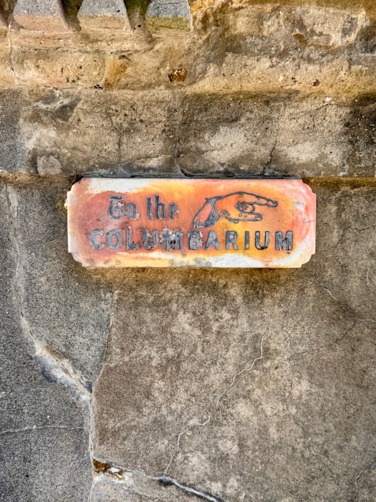 A  rusted white sign on a stone wall reads "To the Columbarium" with a hand pointing to the right. 