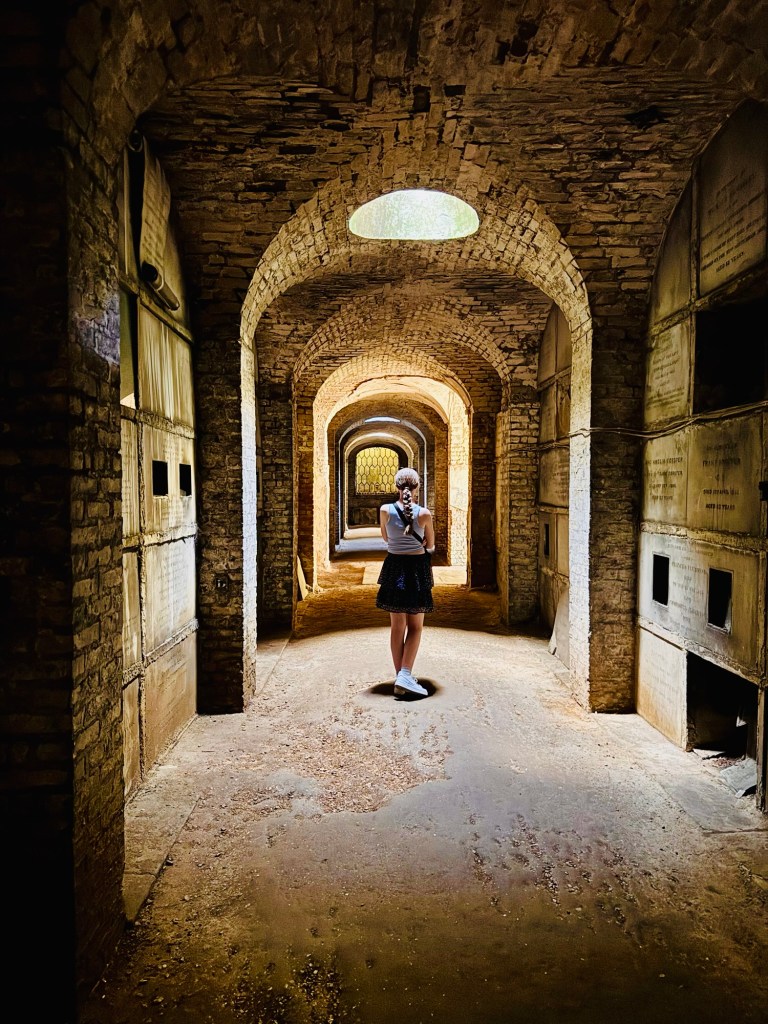 A photo taking inside the catacombs showing a 13 year old girl wearing a light blue top and dark blue skirt standing in a shaft of light coming in from one of the skylights. 